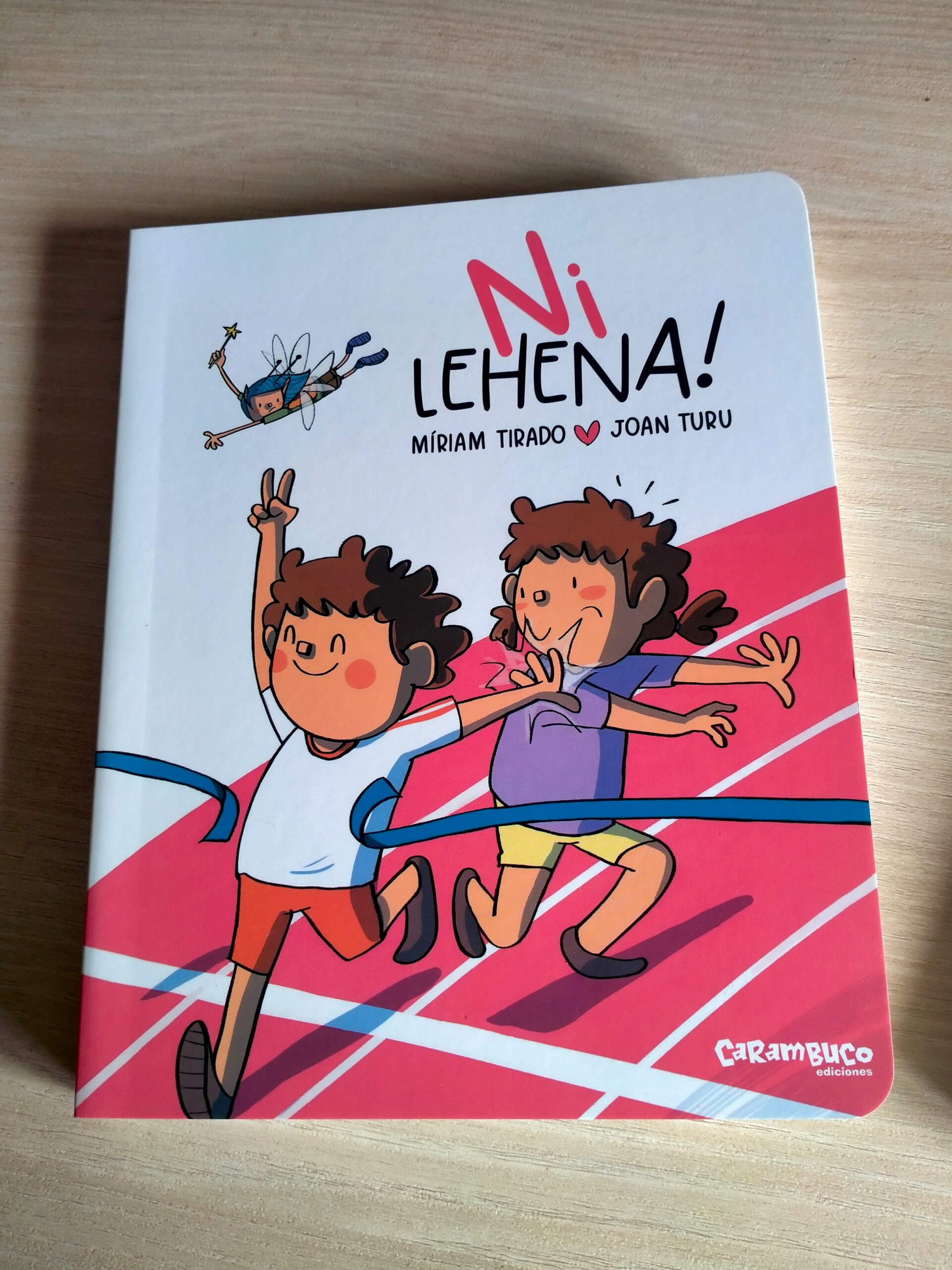 «Ni lehena!» liburuaren azala. Bi haur agertzen dira: mutilak helmuga-zinta bat zeharkatzen ari da korrika eta poz-pozik eta atzean daukan neska eskuarekin aldentzen du. Gainean maitagarri bat dago hegan.