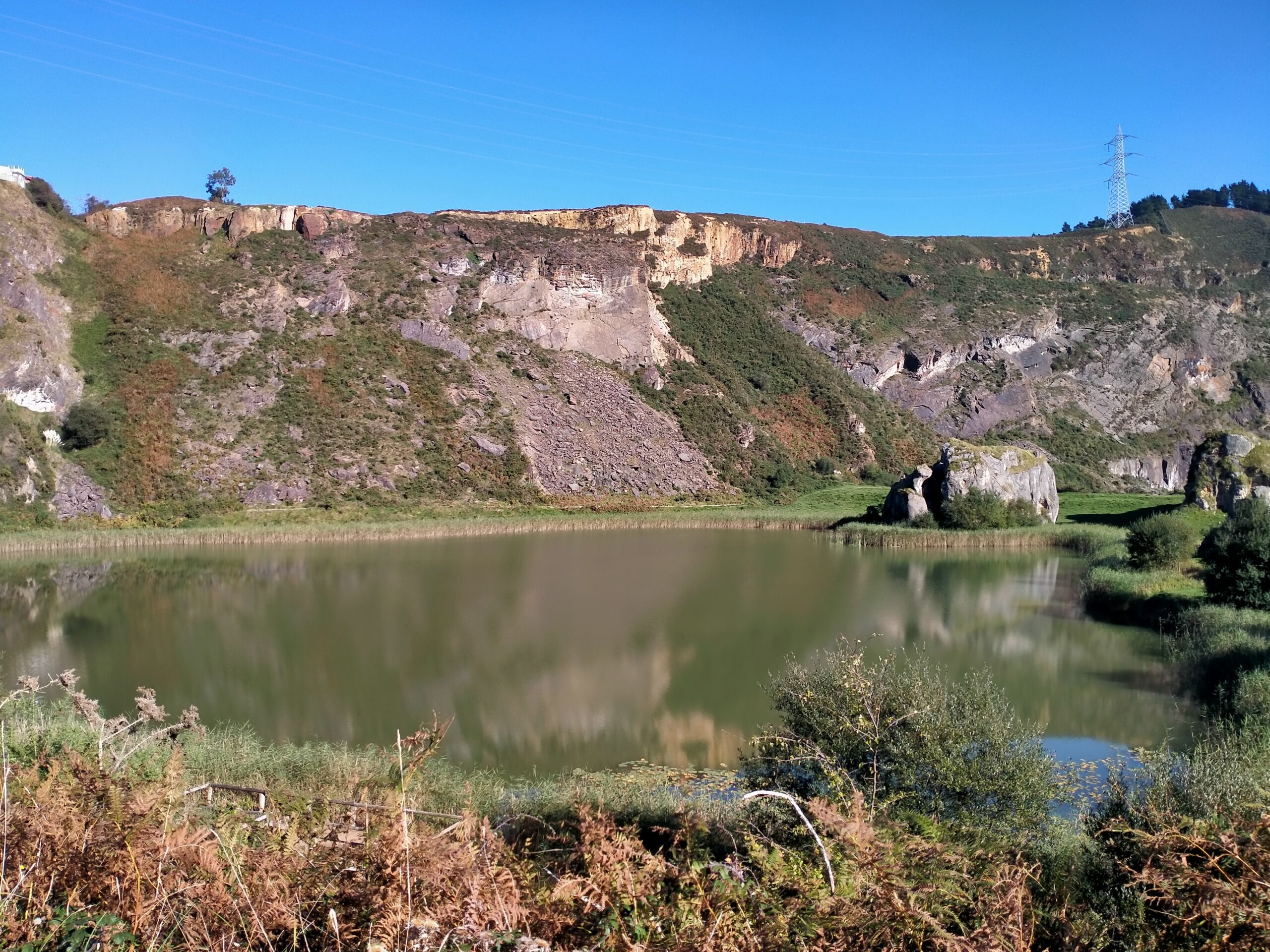 Antiguo pozo minero, ahora convertido en lago. Al fondo se ve una montaña rocosa con algo de vegetación diseminada y un poste eléctrico en lo alto..