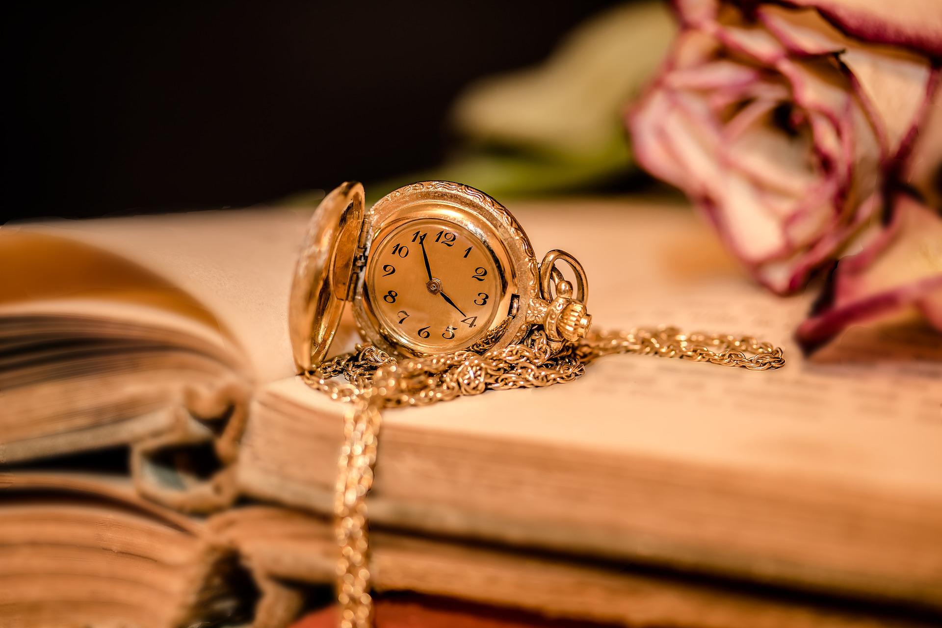 Reloj de bolsillo dorado que marca las cuatro menos cinco, de pie encima de un libro antiguo abierto sobre el que también hay una flor rosa claro. El libro y la flor se ven borrosos, mientras que el reloj es nítido y brillante.